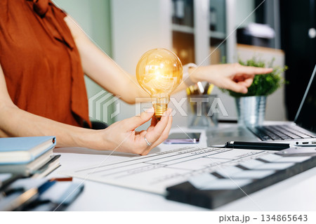 Creative woman holds a glowing lightbulb while using a laptop and clapperboard in modern office. Symbol of inspiration, film production, and innovative ideas. 134865643