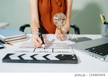 Creative woman holds a glowing lightbulb while using a laptop and clapperboard in modern office. Symbol of inspiration, film production, and innovative ideas. Creative woman holds a glowing lightbulb while using a laptop and clapperboard in modern office. Symbol of inspiration, film production, and innovative ideas. 134865644