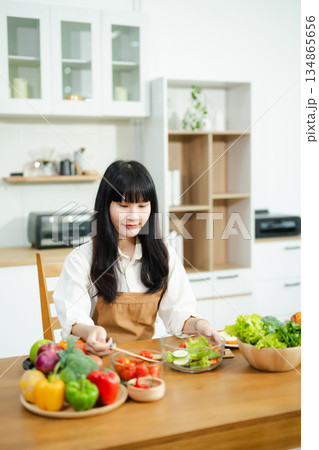 Asian woman prepares healthy salad with fresh vegetables in a modern kitchen. Clean, vibrant, and perfect for nutrition, lifestyle, or home cooking concepts. 134865656