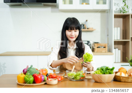 Asian woman prepares healthy salad with fresh vegetables in a modern kitchen. Clean, vibrant, and perfect for nutrition, lifestyle, or home cooking concepts. 134865657