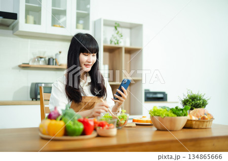 Asian woman checks her phone while holding cherry tomatoes in a modern kitchen. A vibrant healthy cooking scene full of fresh vegetables and lifestyle energy. Asian woman checks her phone while holding cherry tomatoes in a modern kitchen. A vibrant healthy cooking scene full of fresh vegetables and lifestyle energy. 134865666