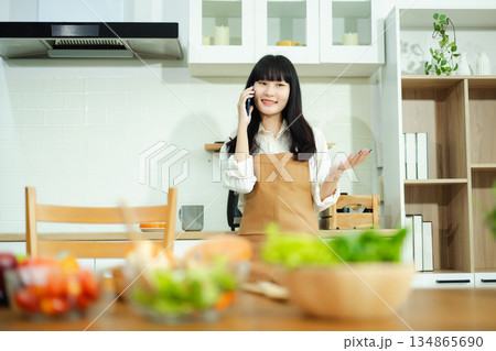 Asian woman chats on the phone and drinks coffee in a cozy kitchen with fresh vegetables. A vibrant lifestyle scene perfect for food, wellness, and communication concepts. Asian woman chats on the phone and drinks coffee in a cozy kitchen with fresh vegetables. A vibrant lifestyle scene perfect for food, wellness, and communication concepts. 134865690