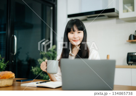 Asian woman working remotely on a laptop and tablet in a cozy kitchen workspace. Perfect for education, freelance Asian woman working remotely on a laptop and tablet in a cozy kitchen workspace. Perfect for education, freelance 134865738