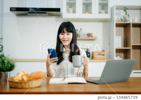 Asian woman working remotely on a laptop and tablet in a cozy kitchen workspace. Perfect for education, freelance Asian woman working remotely on a laptop and tablet in a cozy kitchen workspace. Perfect for education, freelance 134865739