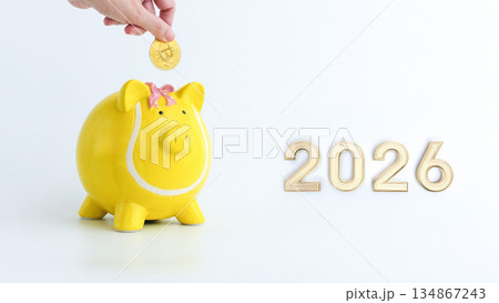 A hand drops a coin into a yellow piggy bank. The year 2026 is displayed next to the piggy bank, symbolizing savings and financial planning for the new year. 134867243
