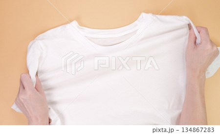 Hands Preparing a Clean White T-Shirt on Beige Background, Ready for Custom Design or Text Hands Preparing a Clean White T-Shirt on Beige Background, Ready for Custom Design or Text 134867283