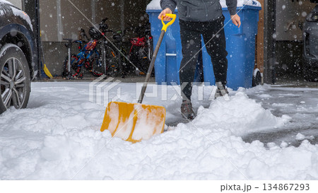 Individual shoveling snow in a residential driveway, surrounded by falling snowflakes and winter scenery, showcasing winter maintenance activity 134867293