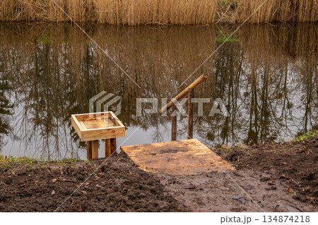 A simple wooden platform stands by a water body, surrounded by grass and trees, reflecting the scenery in the water during the day A simple wooden platform stands by a water body, surrounded by grass and trees, reflecting the scenery in the water during the day 134874218