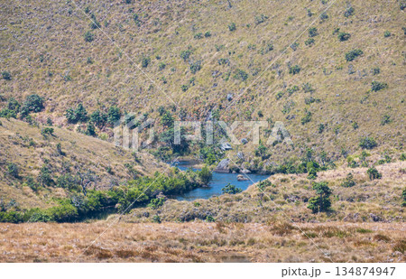 A peaceful river winds through a sunlit grassy valley surrounded by scattered shrubs and rolling hills A peaceful river winds through a sunlit grassy valley surrounded by scattered shrubs and rolling hills 134874947