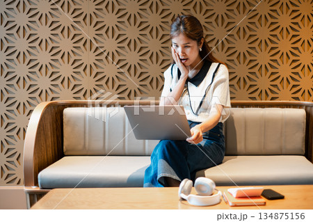 A young woman looks surprised while using a laptop on a modern sofa, set against a stylish geometric wall design. A young woman looks surprised while using a laptop on a modern sofa, set against a stylish geometric wall design. 134875156