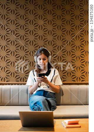 Woman wearing headphones, using smartphone in a modern lounge with geometric wall design. Casual and relaxed atmosphere. 134875160