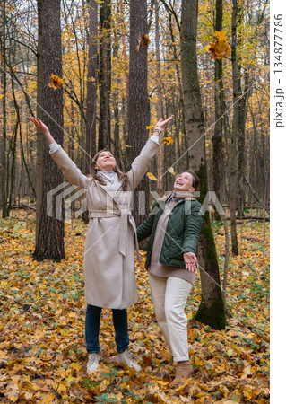 Two women throwing autumn leaves in a forest, enjoying a joyful outdoor moment surrounded by yellow foliage and tall trees Two women throwing autumn leaves in a forest, enjoying a joyful outdoor moment surrounded by yellow foliage and tall trees 134877786