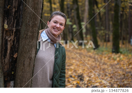 Smiling woman peeks from behind tree in Timiryazevsky Park, Moscow, surrounded by autumn forest with yellow leaves, soft light, friendly, relaxed mood Smiling woman peeks from behind tree in Timiryazevsky Park, Moscow, surrounded by autumn forest with yellow leaves, soft light, friendly, relaxed mood 134877826