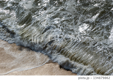 Coastal waves crashing on sandy shore from above, creating frothy foam and textures Coastal waves crashing on sandy shore from above, creating frothy foam and textures 134879862
