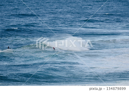 Surfers ride a powerful ocean wave under clear blue sky in a dramatic coastal scene Surfers ride a powerful ocean wave under clear blue sky in a dramatic coastal scene 134879899