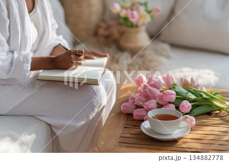 Woman journaling in morning practicing self love with cup of tea and pink tulip flowers on wooden table in cozy living room with soft natural light 134882778