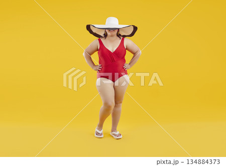 Funny fat woman in swimsuit and big beach hat isolated on studio yellow background. 134884373