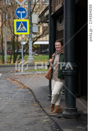 Woman stands near black columns of a historic tram stop pavilion, wearing a green jacket and beige trousers, with pedestrian crossing signs, roadway Woman stands near black columns of a historic tram stop pavilion, wearing a green jacket and beige trousers, with pedestrian crossing signs, roadway 134884898
