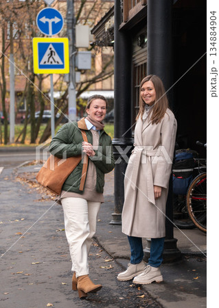 Two women talk and laugh near historic wooden tram stop in Moscow, with wet pavement, tram tracks, street signs, autumn trees in the city background 134884904