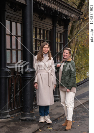 Two women walk past a historic wooden tram stop in Moscow, wearing beige and green coats, smiling, with autumn tones and classic urban surroundings 134884905