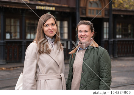 Two women walk past a historic wooden tram stop in Moscow, wearing beige and green coats, smiling, with autumn tones and classic urban surroundings 134884909