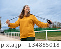 A woman in a yellow hoodie holds a jump rope and smiles against a sky background. 134885018