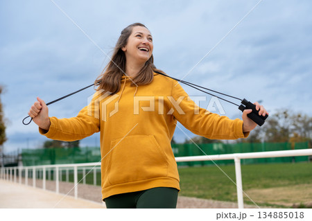 A woman in a yellow hoodie holds a jump rope and smiles against a sky background. 134885018