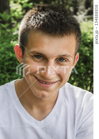 Close-up portrait of a smiling young man wearing a white T-shirt, looking at the camera against a natural green background. Happy teenager with short hair and blue eyes. Close-up portrait of a smiling young man wearing a white T-shirt, looking at the camera against a natural green background. Happy teenager with short hair and blue eyes. 134888938