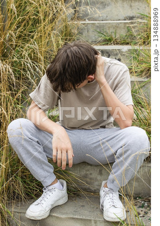 A lonely teenager sits on concrete steps among dry grass, head down, reflecting sadness, depression, and youth mental health struggles. A lonely teenager sits on concrete steps among dry grass, head down, reflecting sadness, depression, and youth mental health struggles. 134888969