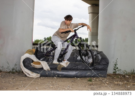 Teenage boy posing with a bicycle on an old sofa under a bridge. Urban lifestyle scene with youth, freedom and creativity in an industrial outdoor environment.  134888973