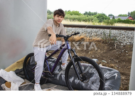 Teenage boy posing with a bicycle on an old sofa under a bridge. Urban lifestyle scene with youth, freedom and creativity in an industrial outdoor environment. Teenage boy posing with a bicycle on an old sofa under a bridge. Urban lifestyle scene with youth, freedom and creativity in an industrial outdoor environment. 134888979