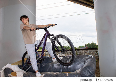 Teenage boy posing with a bicycle on an old sofa under a bridge. Urban lifestyle scene with youth, freedom and creativity in an industrial outdoor environment. Teenage boy posing with a bicycle on an old sofa under a bridge. Urban lifestyle scene with youth, freedom and creativity in an industrial outdoor environment. 134888980