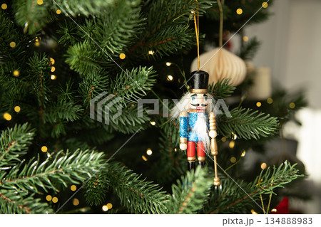 Traditional wooden nutcracker Christmas ornament hanging on a decorated Christmas tree with warm lights and golden baubles. Festive holiday atmosphere with cozy winter mood. 134888983