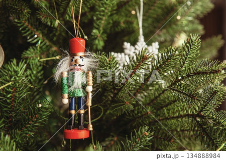 Traditional wooden nutcracker Christmas ornament hanging on a decorated Christmas tree with warm lights and golden baubles. Festive holiday atmosphere with cozy winter mood. 134888984