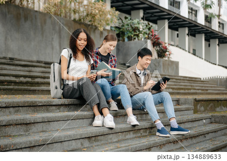 Three young college students is reading a book while relaxing sitting on grass in a campus park with her friends. Three young college students is reading a book while relaxing sitting on grass in a campus park with her friends. 134889633