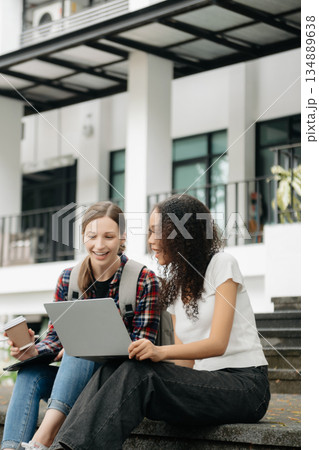 Three young college students is reading a book while relaxing sitting on grass in a campus park with her friends. Three young college students is reading a book while relaxing sitting on grass in a campus park with her friends. 134889638