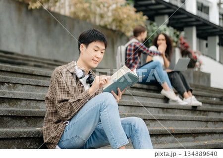 Three young college students is reading a book while relaxing sitting on grass in a campus park with her friends. 134889640