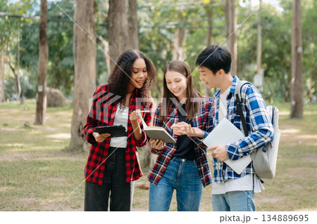 Three young college students is reading a book while relaxing sitting on grass in a campus park with her friends. 134889695