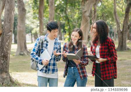 Three young college students is reading a book while relaxing sitting on grass in a campus park with her friends. Three young college students is reading a book while relaxing sitting on grass in a campus park with her friends. 134889696