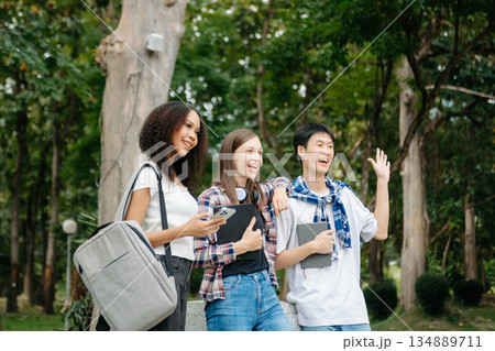 Three young college students is reading a book while relaxing sitting on grass in a campus park with her friends. 134889711