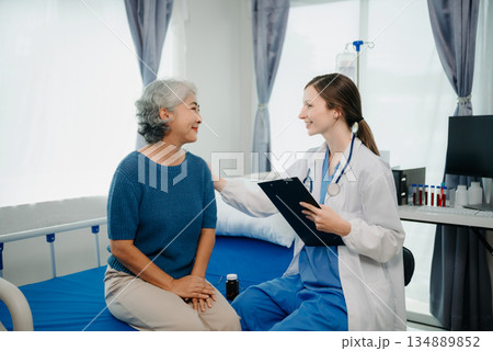 Doctor in white suit take notes while discussing and Asian elderly, woman patient who lying on bed with receiving saline solution in hospital or clinic. Doctor in white suit take notes while discussing and Asian elderly, woman patient who lying on bed with receiving saline solution in hospital or clinic. 134889852