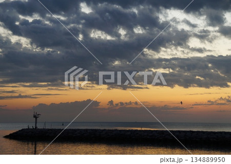The first rays of sun paint the gray sky orange. The gray and orange sky is reflected in the sea, against the backdrop of a stone pier  134889950