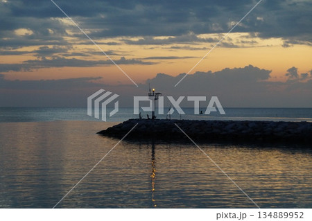 The first rays of sun paint the gray sky orange. The gray and orange sky is reflected in the sea, against the backdrop of a stone pier.The dawn is breaking over the horizon. 134889952