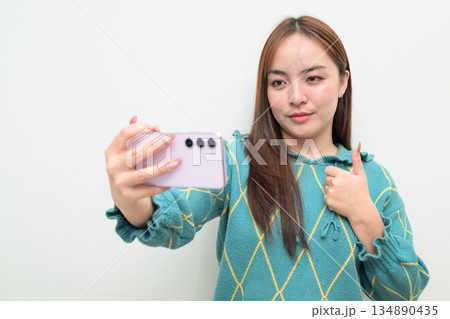 Portrait of a young Asian woman against white background using phone taking selfie 134890435