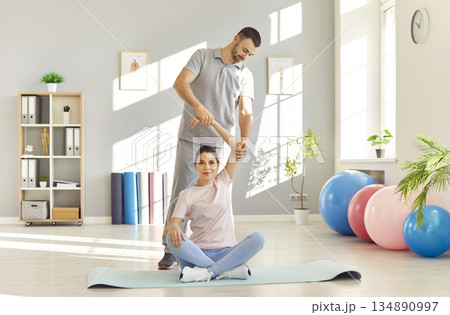 Physical therapist helping woman in doing exercises sitting on mat in rehabilitation center. Physical therapist helping woman in doing exercises sitting on mat in rehabilitation center. 134890997