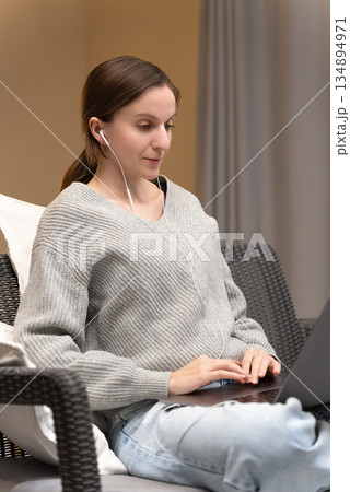 Young Caucasian woman in gray sweater sitting indoors with earphones, work from home office. Student use pc and internet e-learning remotely, watching webinar or tutorial studying online on laptop 134894971