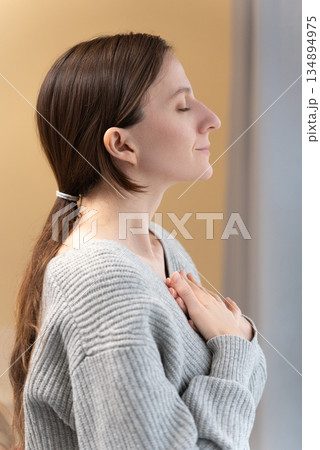 Portrait serene peaceful young Caucasian woman with closed eyes practicing deep breathing, hands on chest, feeling calm and peaceful, demonstrating stress relief, inner peace and mindfulness concept 134894975