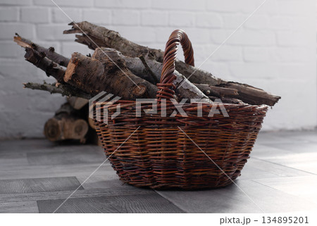 Wicker Basket with Firewood near Fireplace in a cozy home interior. Stacked logs in the background. Winter heating rustic lifestyle concept. Wicker Basket with Firewood near Fireplace in a cozy home interior. Stacked logs in the background. Winter heating rustic lifestyle concept. 134895201