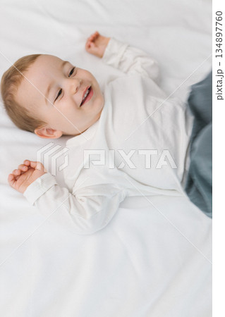 Cozy portrait of a happy baby lying on a soft white bed at home, smiling in natural light. A warm and cozy childhood moment filled with joy, innocence, comfort, and gentle family emotion. 134897760
