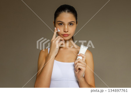Young woman applying face cream while holding a cosmetic tube, demonstrating a hydrating skincare step on a clean studio background. Young woman applying face cream while holding a cosmetic tube, demonstrating a hydrating skincare step on a clean studio background. 134897761